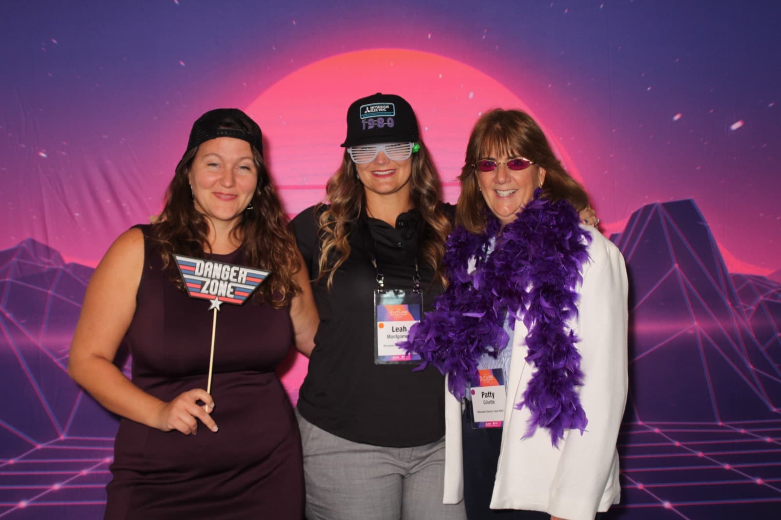 Three colleagues in matching event hats and lanyards smiling at a corporate event photo booth.