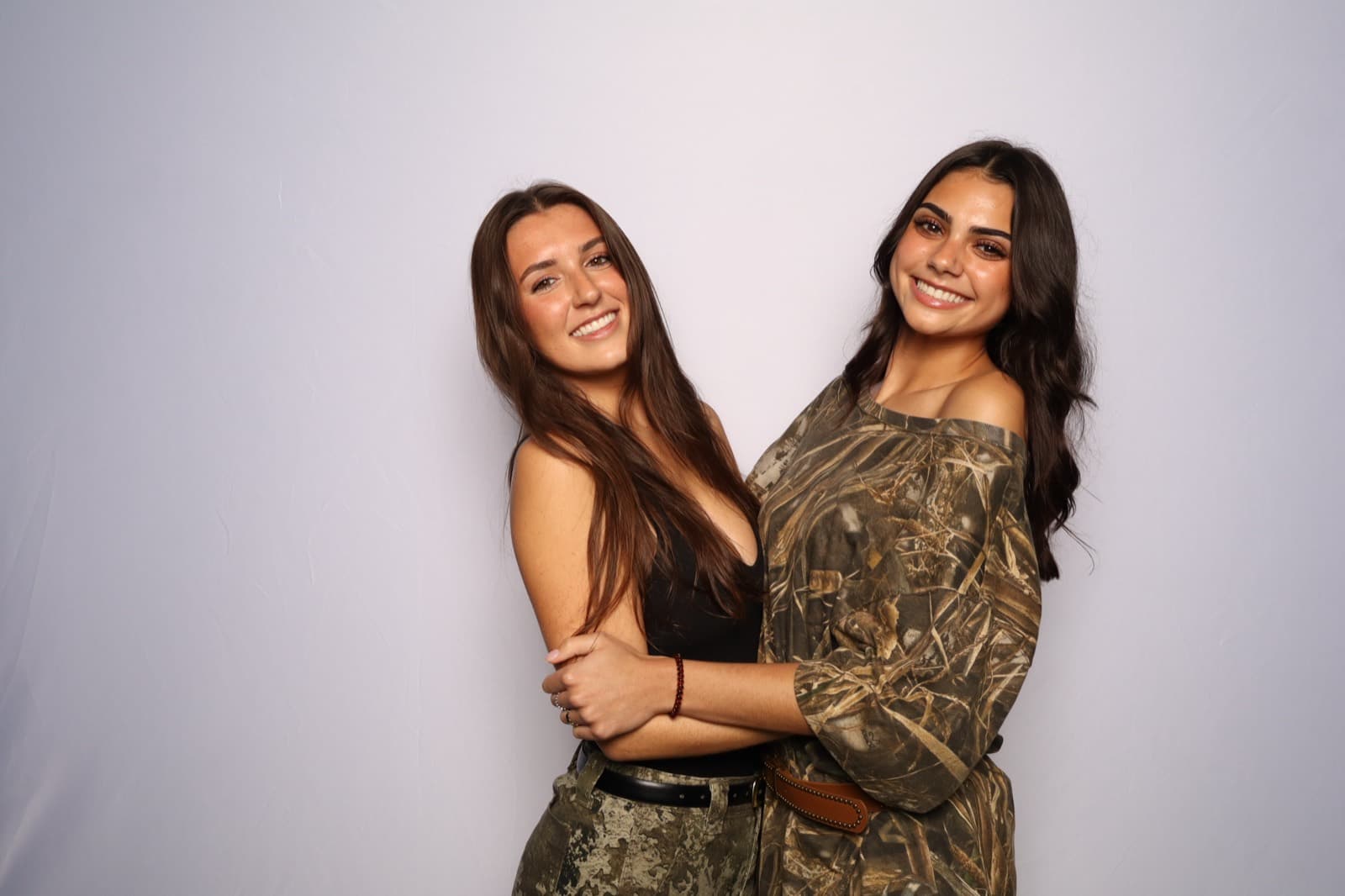 Two friends in camo and black hugging and smiling in front of a pale studio wall.