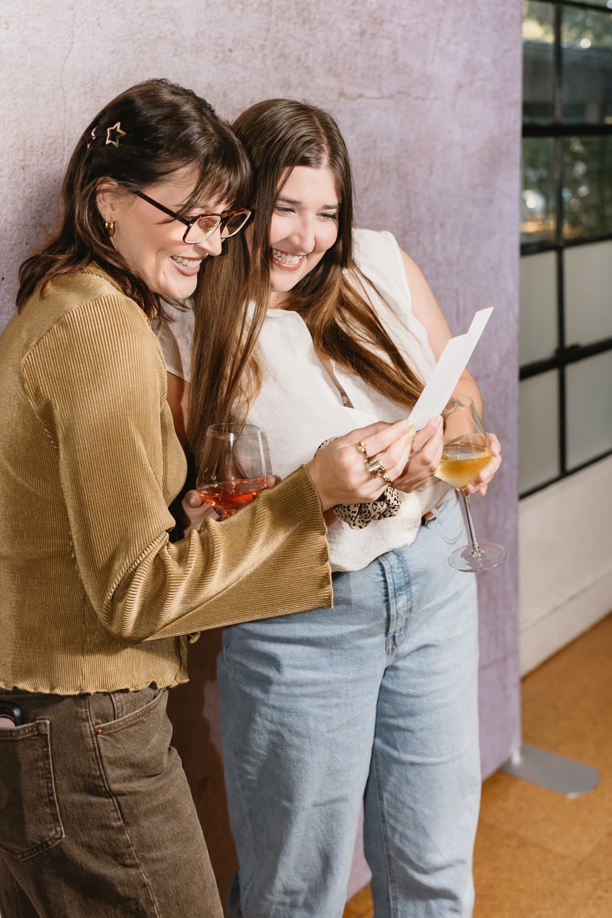 Two friends leaning together and laughing at a photo strip, wine glasses in hand, against a lilac wall.