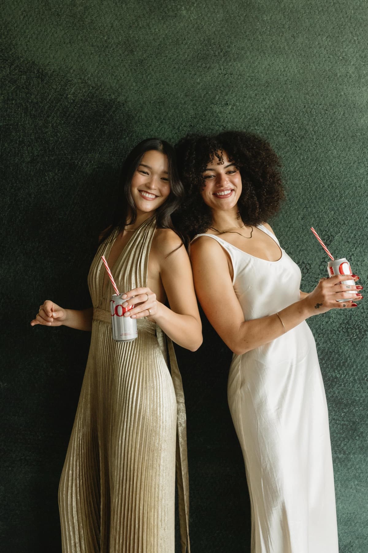 Two women in gold and ivory gowns posing back to back with drinks against a dark green backdrop.