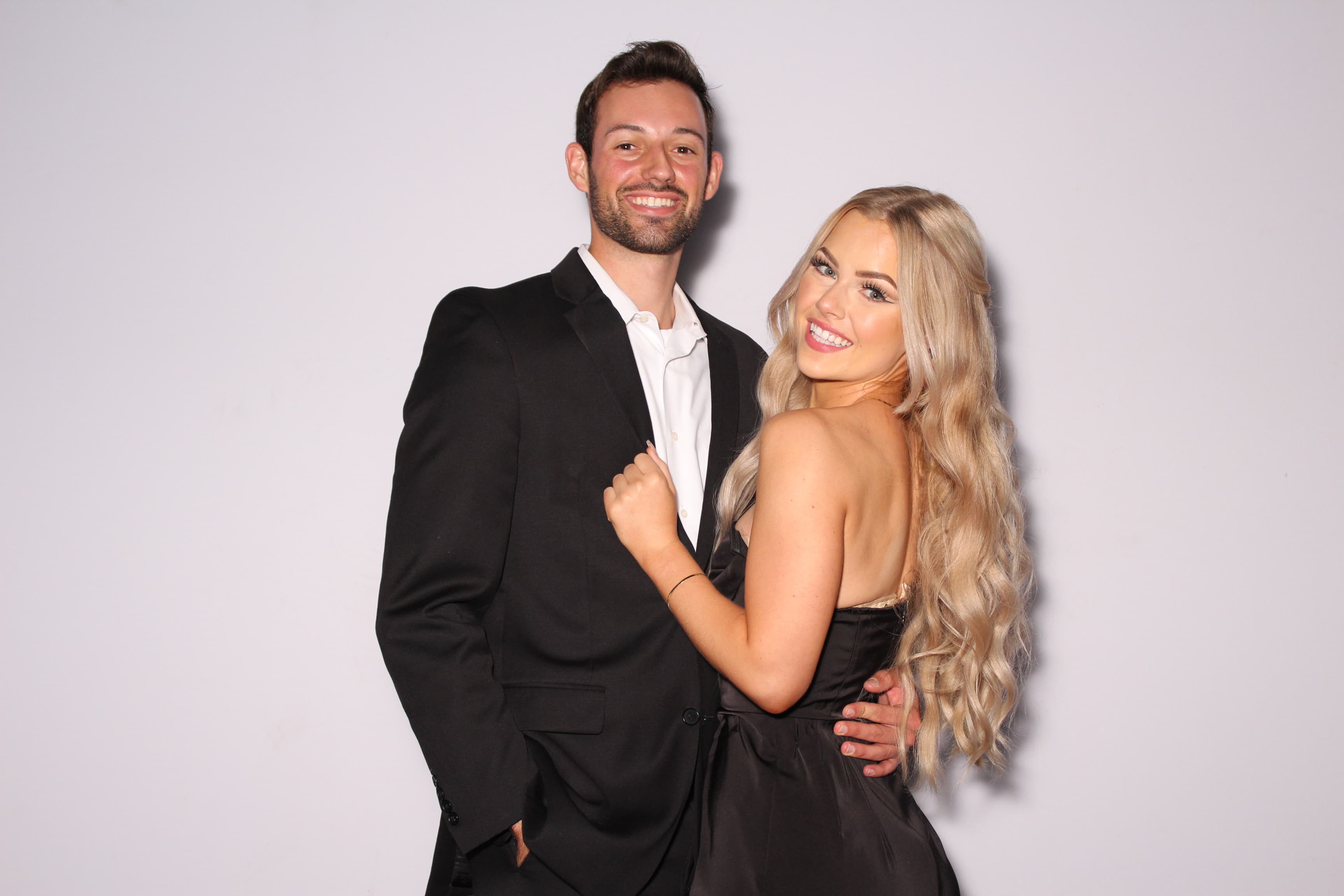 A couple in black formalwear smiling close together in front of a plain white backdrop.