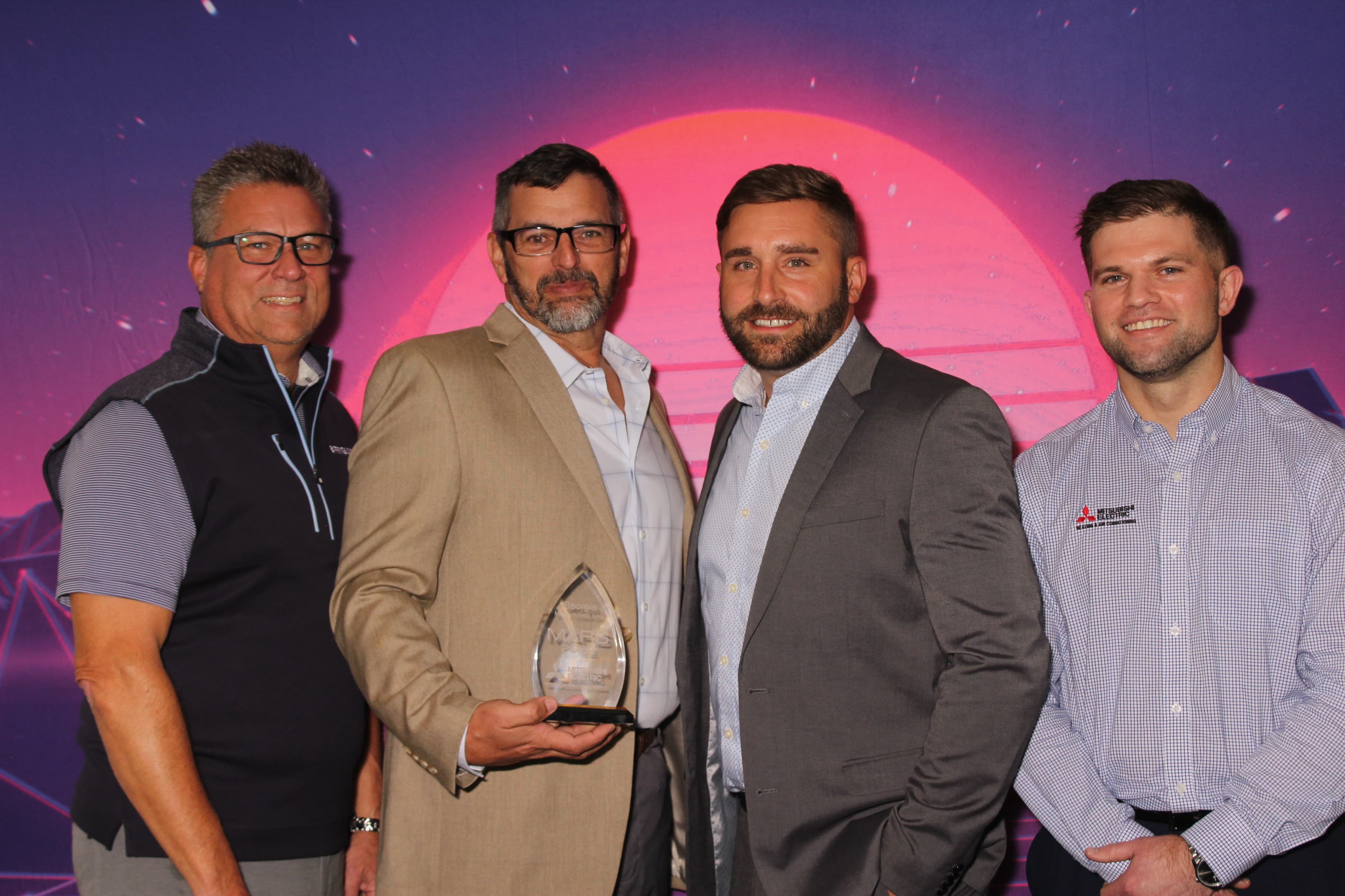 Four colleagues posing with a glass award trophy in front of a retro sunset-themed backdrop at a corporate event.