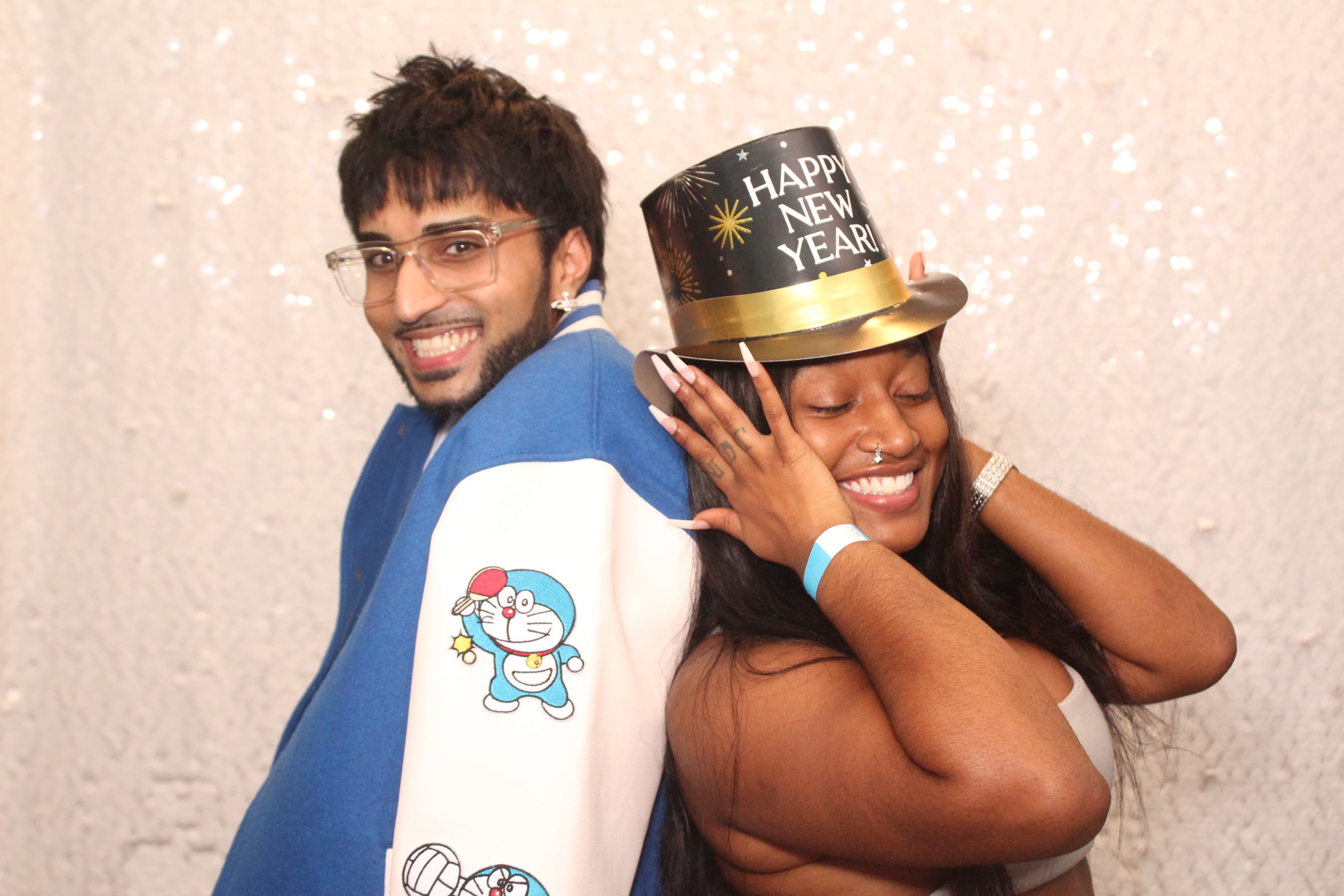 Two friends celebrating New Year’s Eve, one wearing a ‘Happy New Year’ top hat, against a sparkling sequin backdrop.