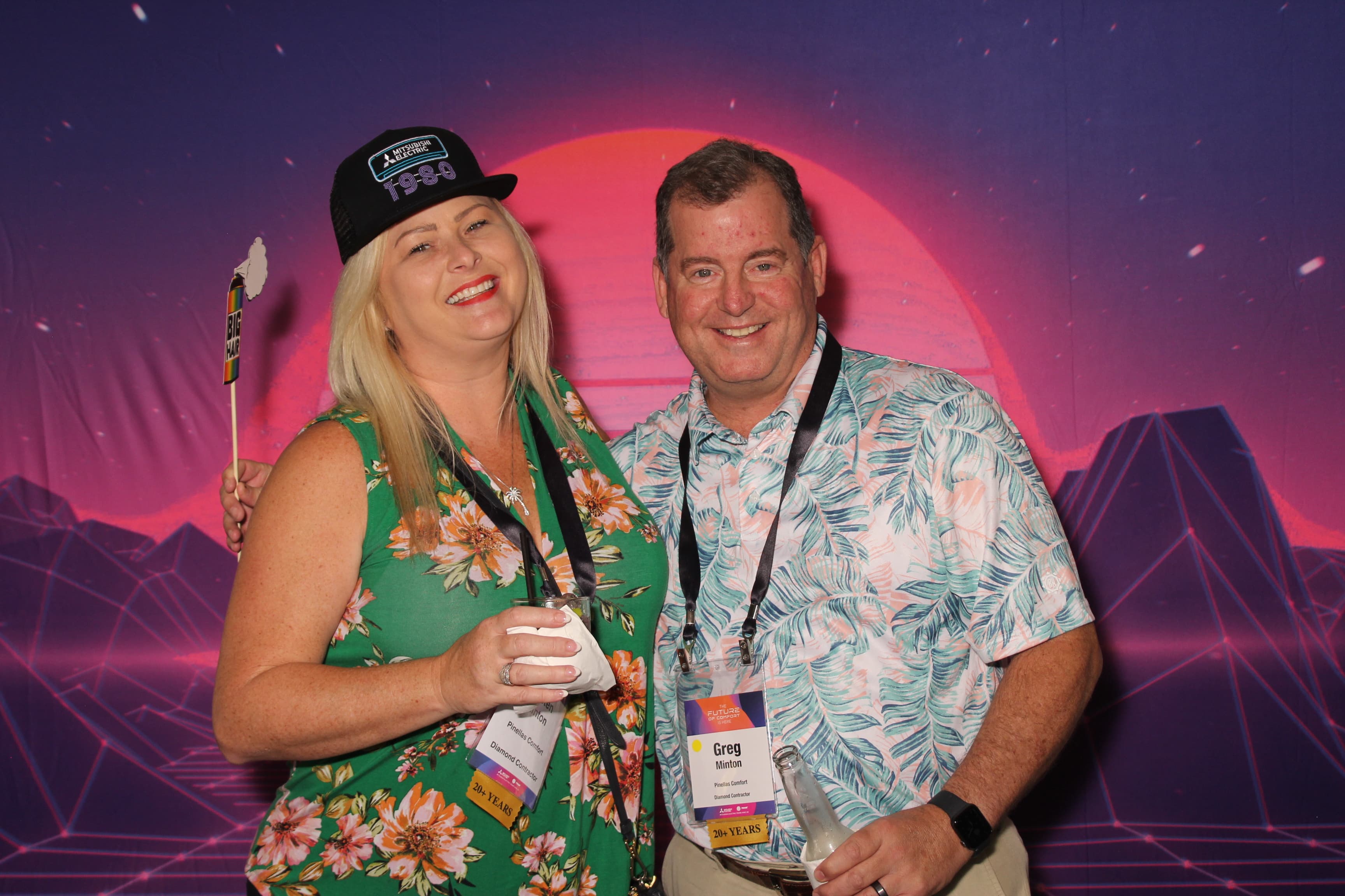 A couple in festive tropical-print outfits posing with a feather prop in front of a retro sunset backdrop at a conference.