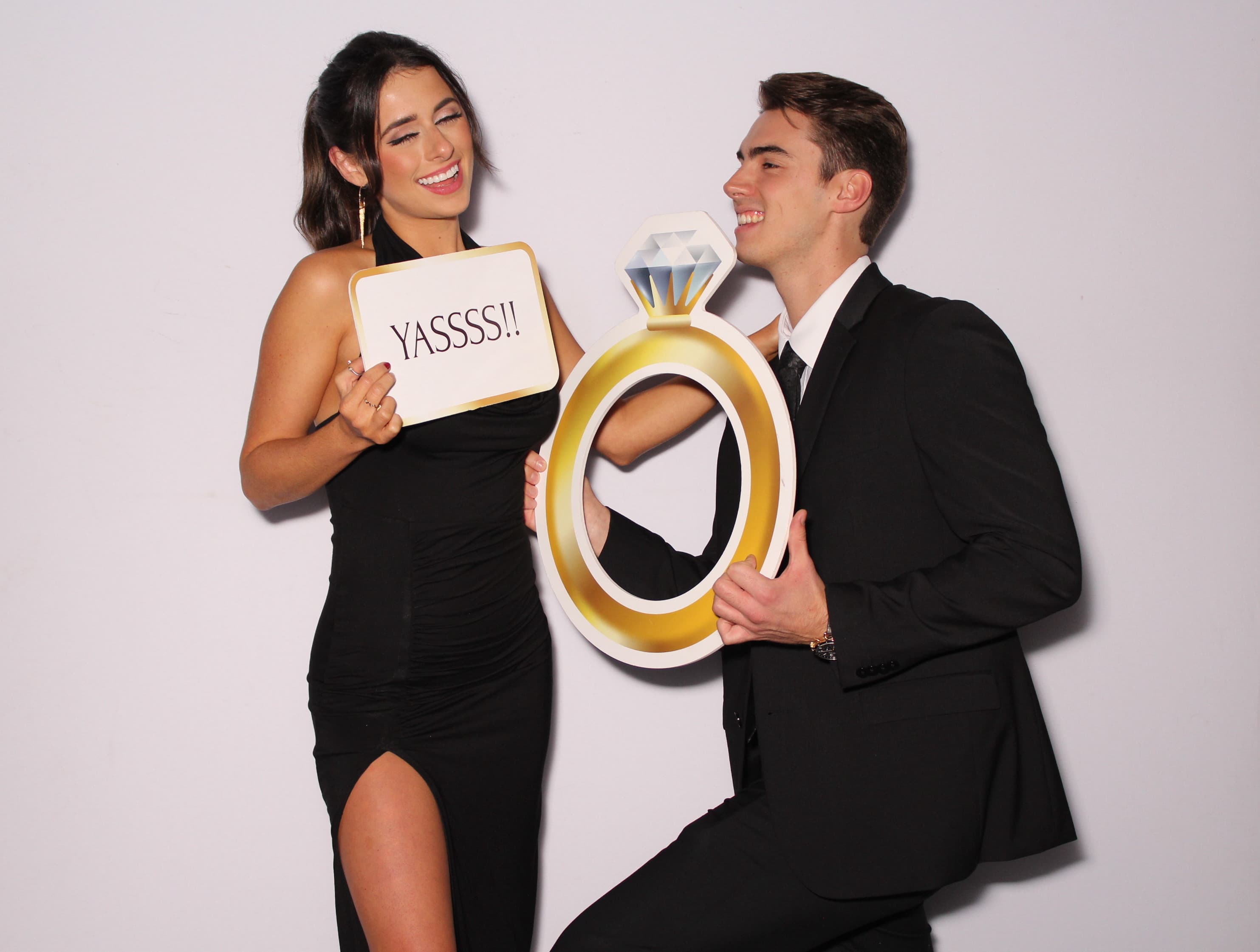 A newly engaged couple posing with a giant engagement-ring cutout and a 'Yassss!' sign.