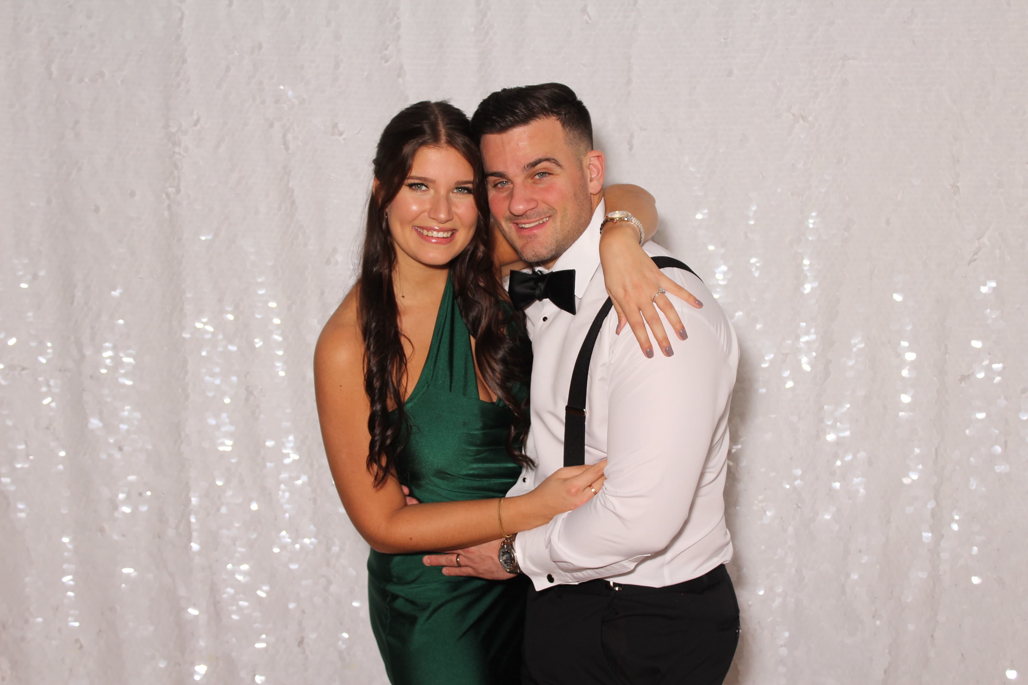 A couple in green satin and black tie embracing in front of a shimmering white sequin backdrop.