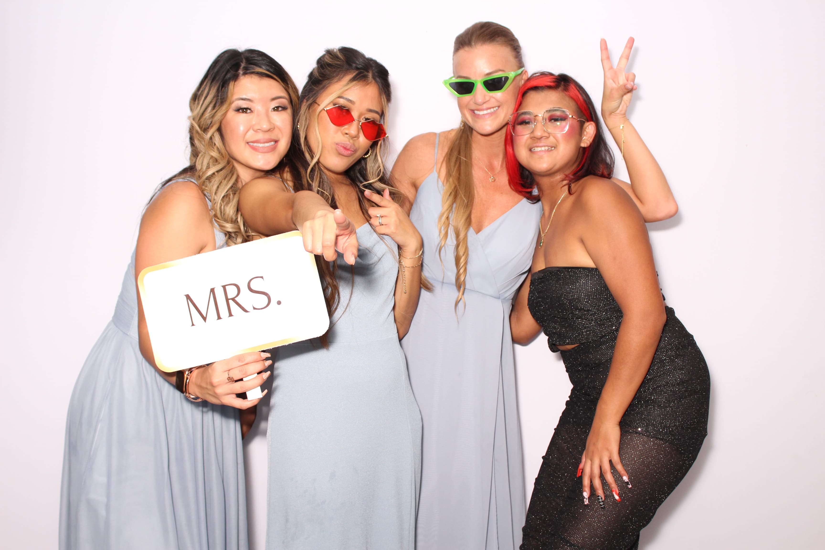 Four bridesmaids in dusty-blue dresses and party sunglasses posing with a ‘MRS.’ sign.