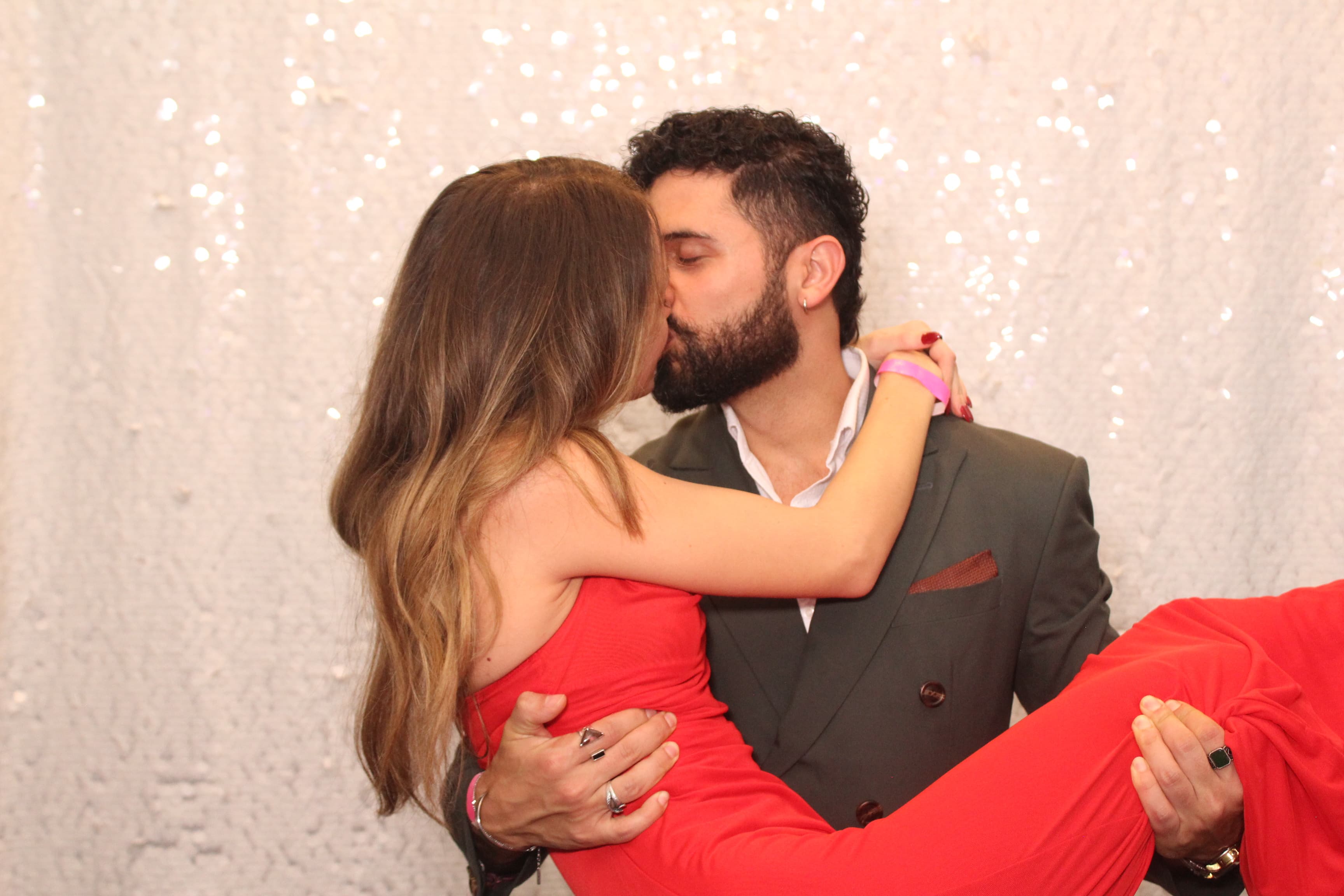 A man lifting a woman in a red dress into his arms for a kiss against a sparkling sequin backdrop.