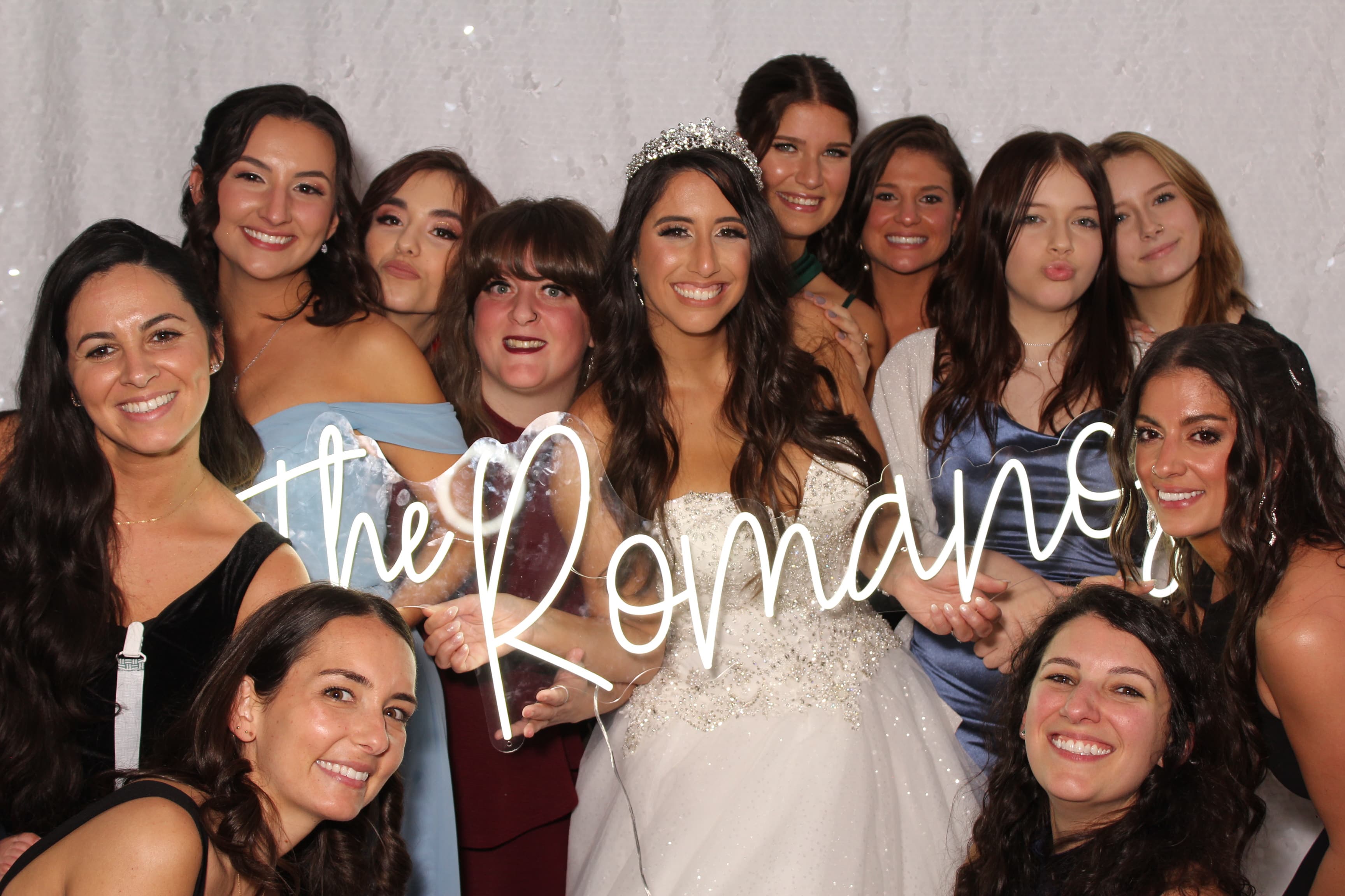 A bride surrounded by her bridal party, all smiling together behind a glowing ‘The Romanos’ neon sign.