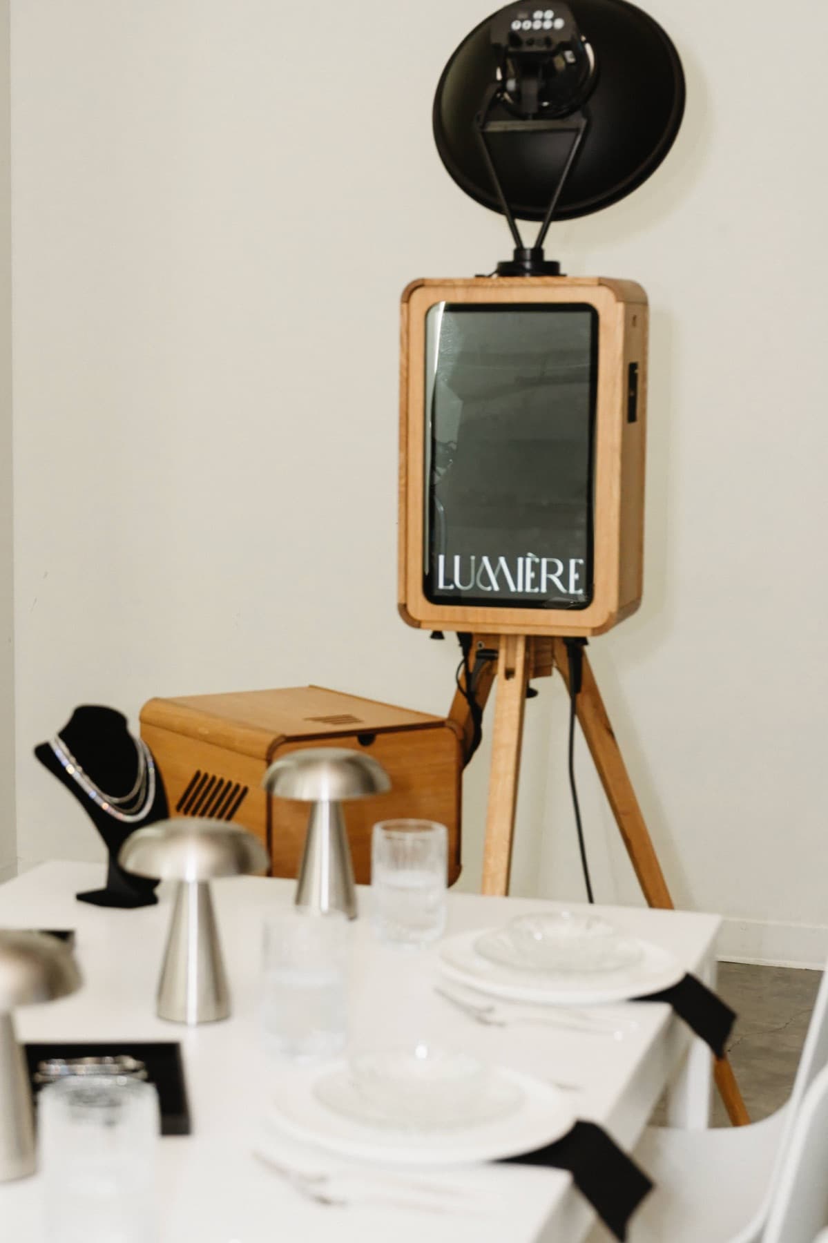 The MEMORI open-air photo booth on its oak tripod beside an elegant place setting, its screen showing the Lumière welcome display.