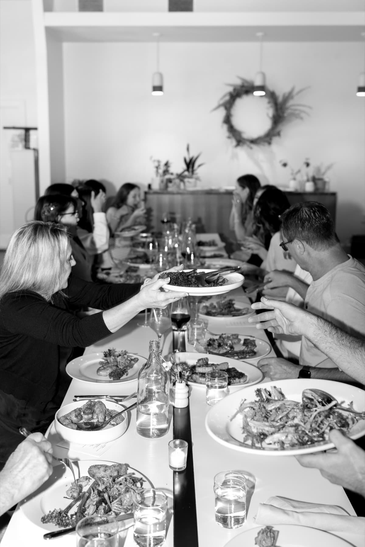 Guests passing plates around a long candlelit dinner table in black and white.