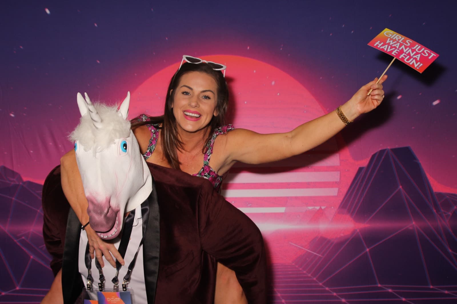 A guest waving a sign prop while riding piggyback on a coworker in a unicorn mask.