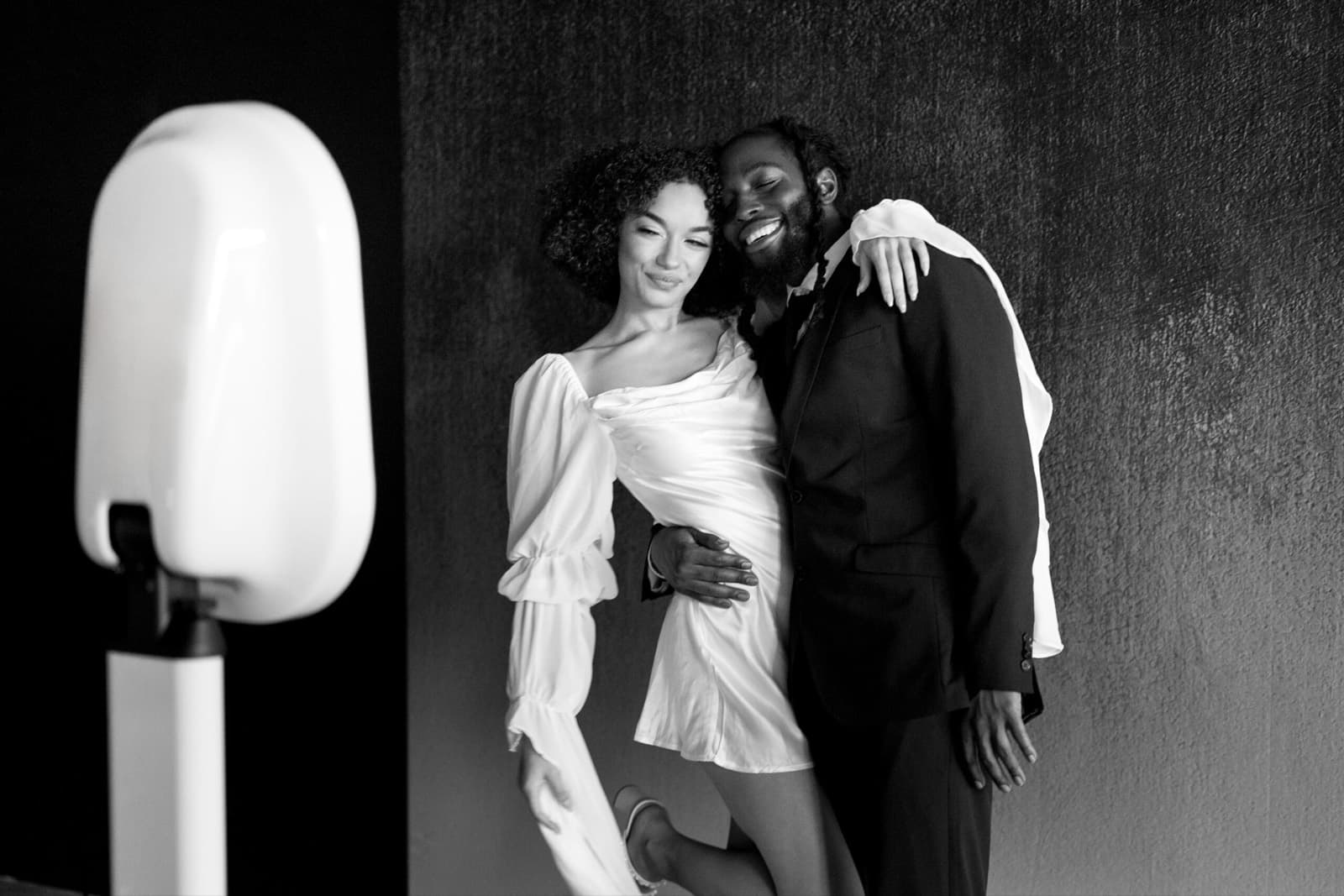 A couple posing beside the MEMORI booth in front of a dark textured wall, in black and white.