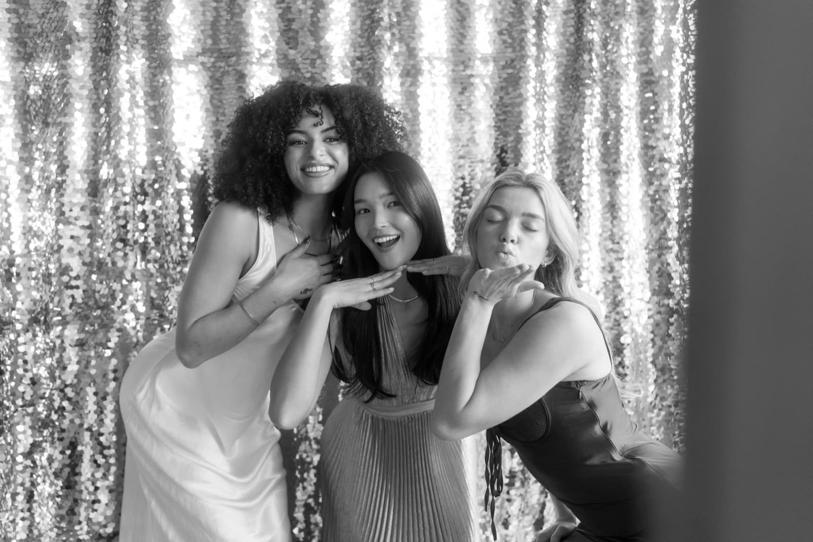 Three friends blowing kisses in front of a silver sequin backdrop, in black and white.