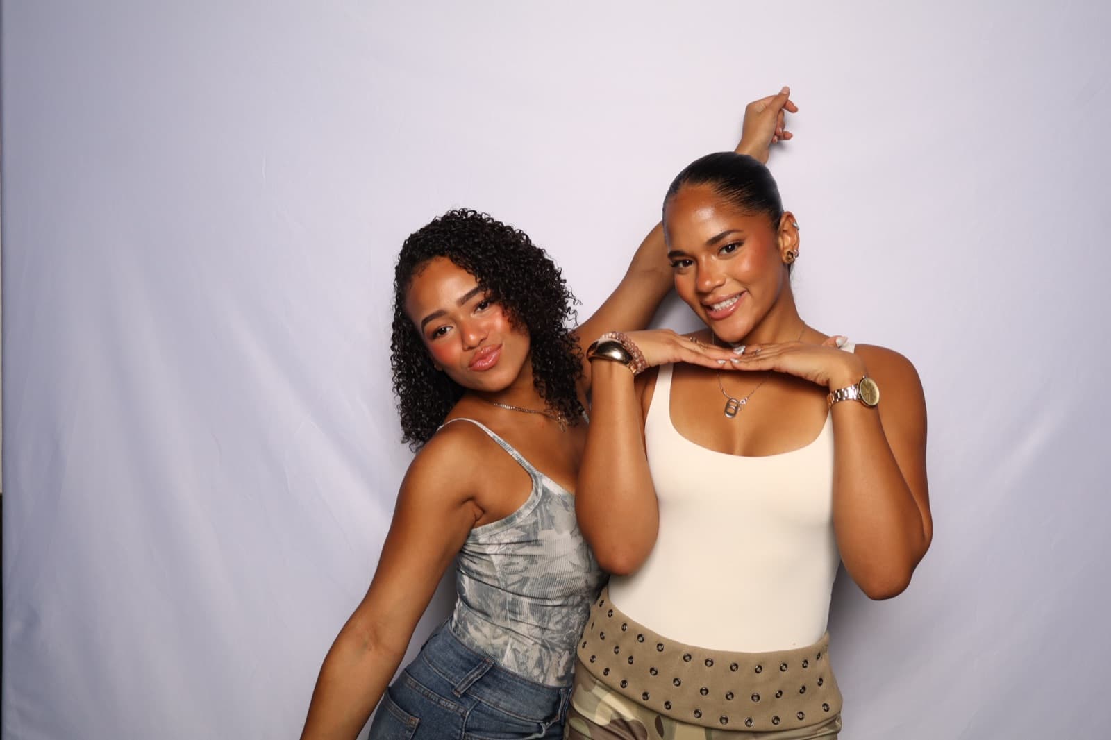 Two friends posing playfully in front of a white backdrop, one resting her hands under the other's chin.