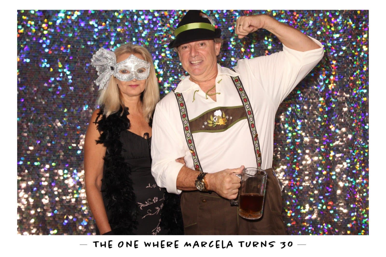 A couple in costume, one in lederhosen raising a beer stein, on a rainbow sequin backdrop at Marcela's 30th birthday.