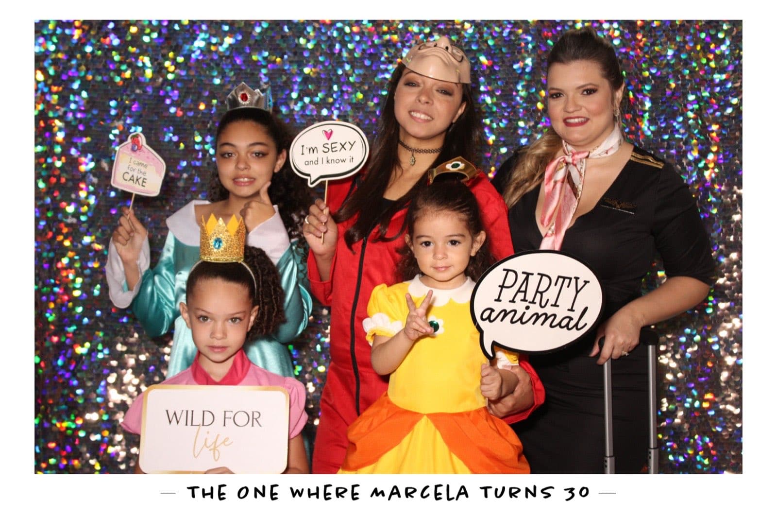 A family in costume with 'Party Animal' and 'Wild for Life' signs on a rainbow sequin backdrop at Marcela's 30th birthday.