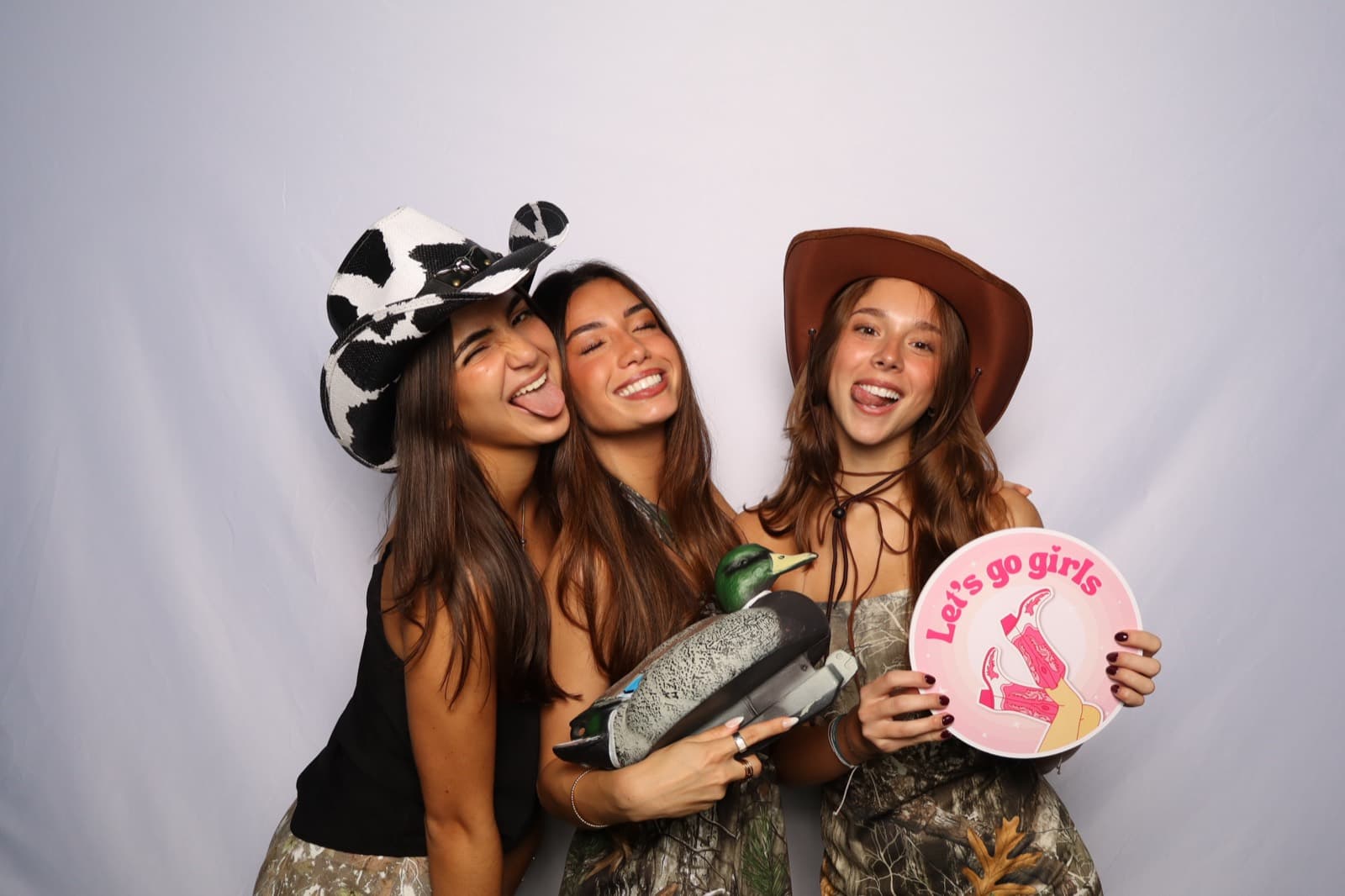 Three friends in cowboy hats laughing with a duck decoy and a 'Let's go girls' sign at the MEMORI booth.
