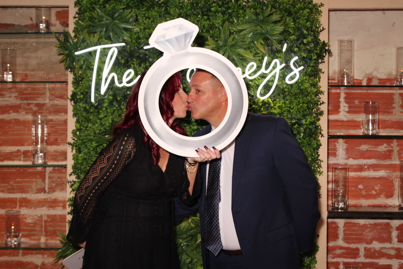 A couple kissing behind a giant ring prop in front of a hedge wall with a custom neon name sign.