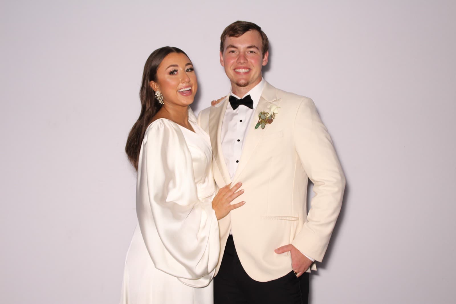 A bride in a satin gown and a groom in a white tuxedo jacket posing at the MEMORI booth.